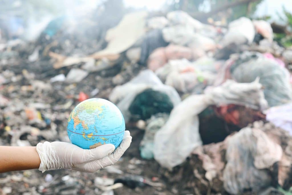 A gloved hand holding a small globe in front of a large pile of plastic waste, symbolizing global plastic pollution.